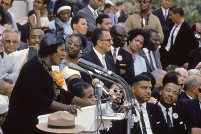 A Black woman is singing in front of several microphones as mostly Black people sit and listen.