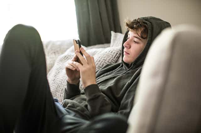 Sitting on a sofa, a teenage boy wearing a hoodie solemnly looks at his smartphone.