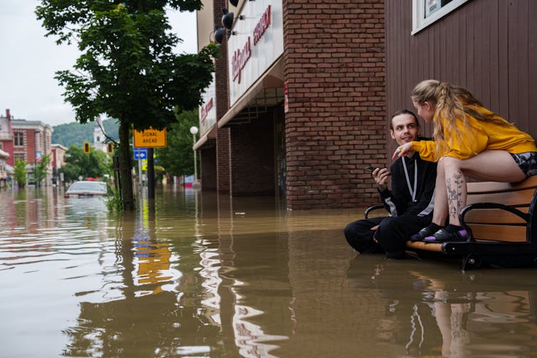 A man and woman sit on a park bench with water up to the man's knees. The woman is sitting on the chair back. A car in the street is flooded up to the roof.