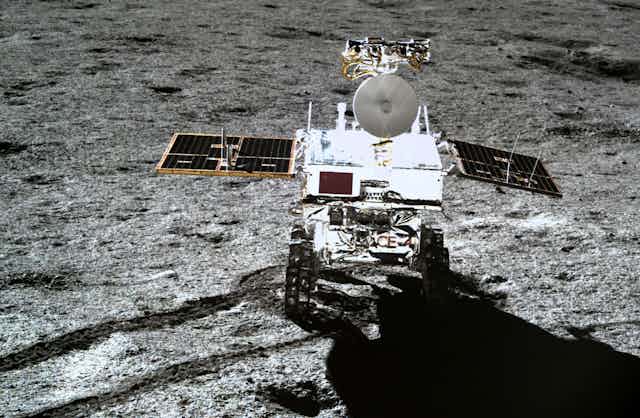 A shiny rover with two solar panels jutting out on either side on a grey rocky surface