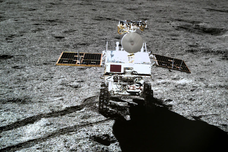 A shiny rover with two solar panels jutting out on either side on a grey rocky surface