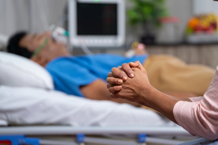 Man lying in hospital bed with oxygen mask, holding hands of female friend or relative