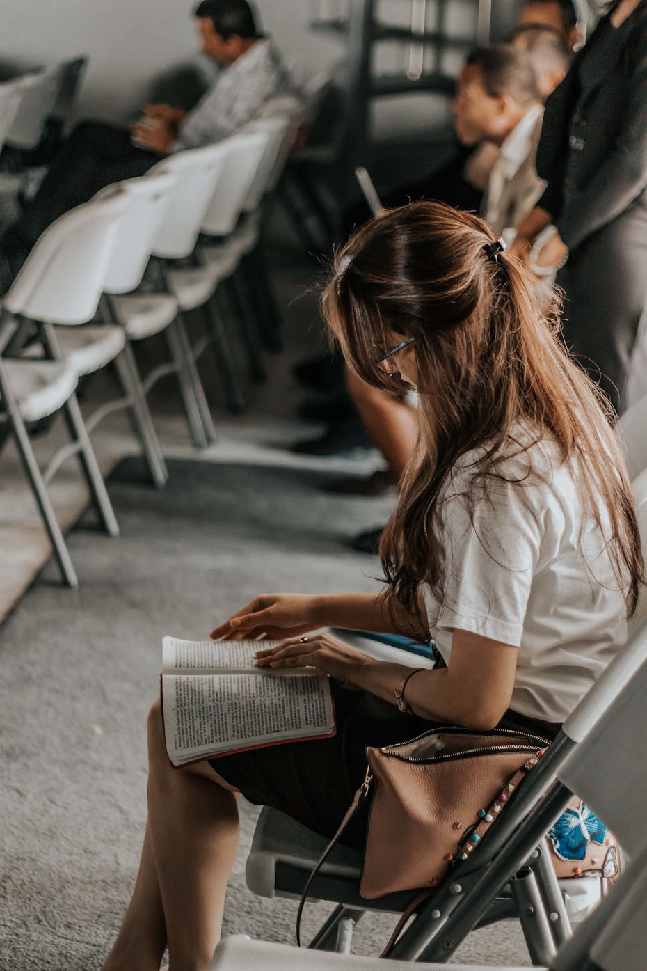 A woman sits at the back of a room reading a book.