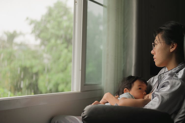 A woman breastfeeds her child while staring out the window sadly.