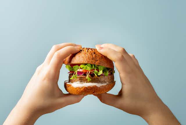 Two hands holding a burger bun with plant-based patty and vegetables, sauce.
