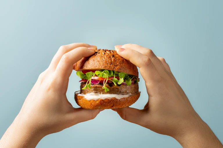 Two hands holding a burger bun with plant-based patty and vegetables, sauce.