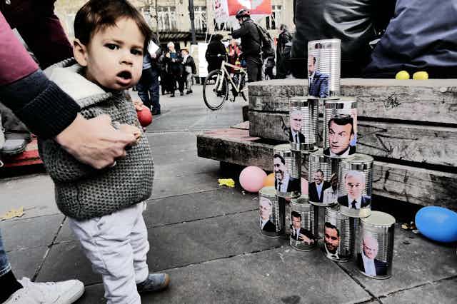 Un niño delante de un montón de latas con efigies de políticos.