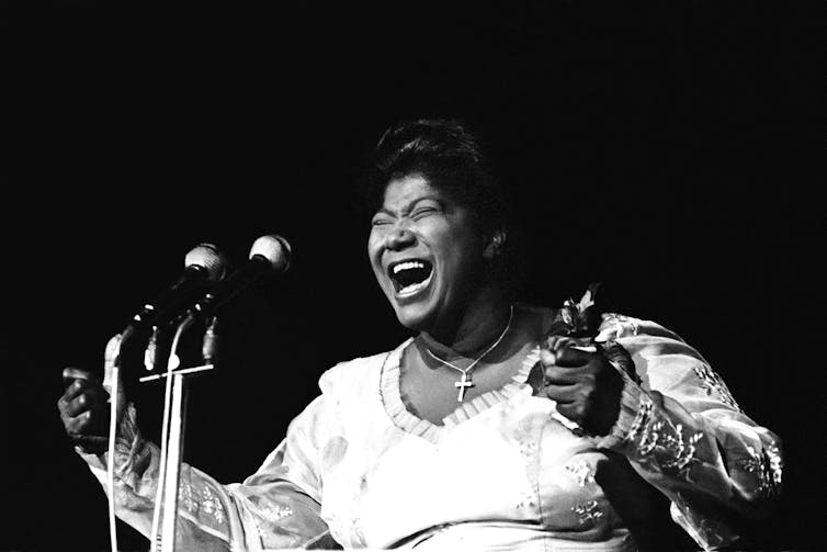 A Black woman dressed in a white gown gestures with her hands as she sings behind several microphones.