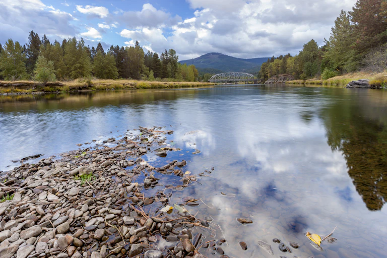 A view of a shallow part of a river with a bridge and mountains in the background. Pine forests line the banks. The image is taken near Cataldo, a few miles from the fire smelter.