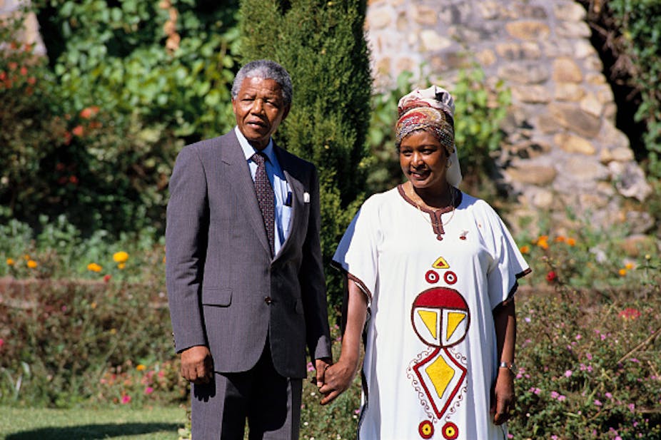 A man wearing a suit holds hands with a woman wearing long African dress and headscarf.