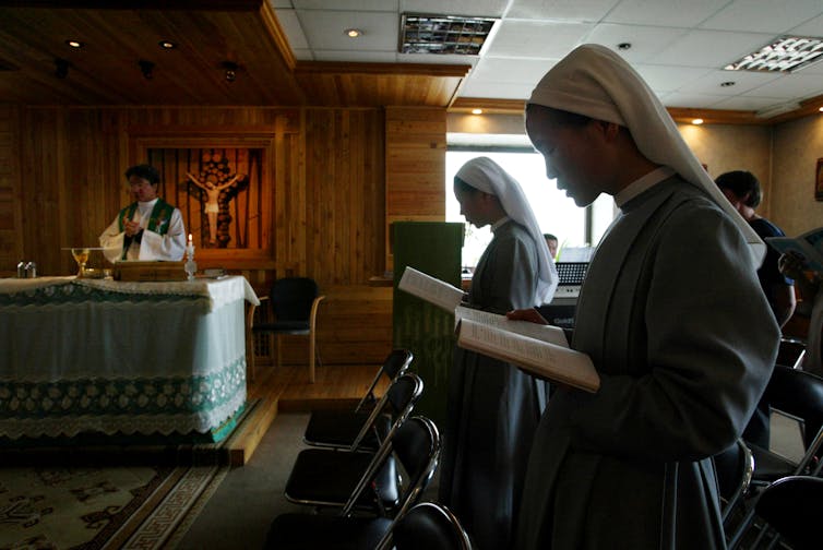 A priest leads a service while worshippers, including two nuns, stand with prayer books and heads bowed.