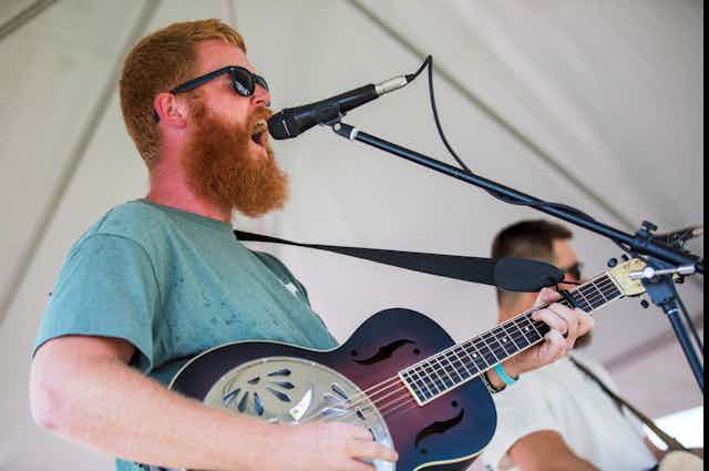 Un homme roux et barbu chante dans un micro et joue de la guitare