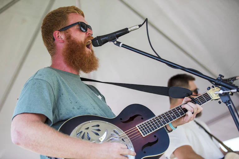 Un homme roux et barbu chante dans un micro et joue de la guitare