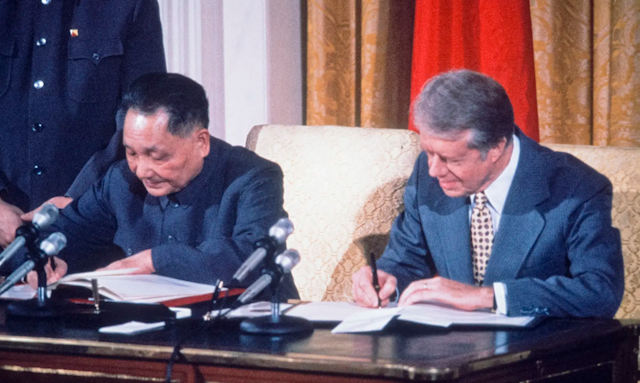 Two men sit at a wooden desk and sign papers