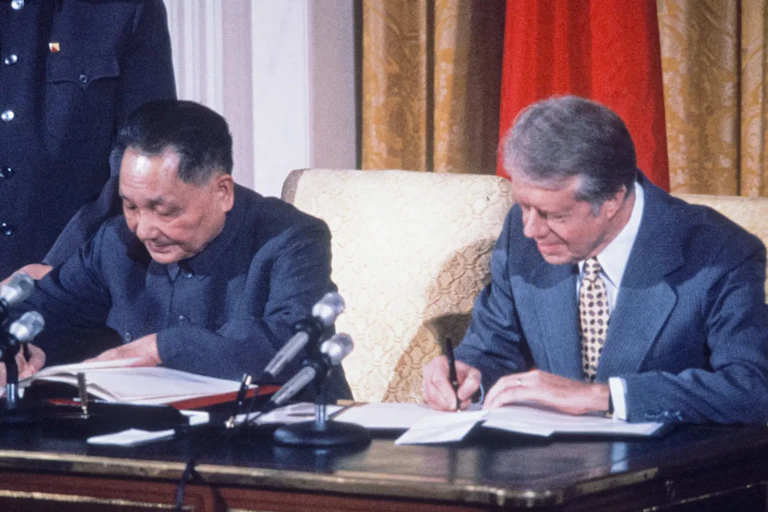 Two men sit at a wooden desk and sign papers