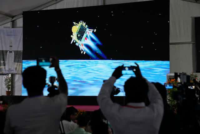 People hold phones up to film a large screen depicting the lander (yellow) as it moves toward the lunar surface (shown as blue).