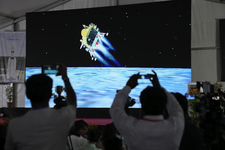 People hold phones up to film a large screen depicting the lander (yellow) as it moves toward the lunar surface (shown as blue).