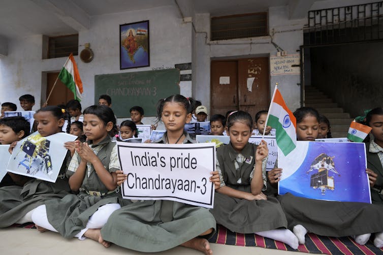 Young people sitting on a rug in a classroom hold flags and signs reading