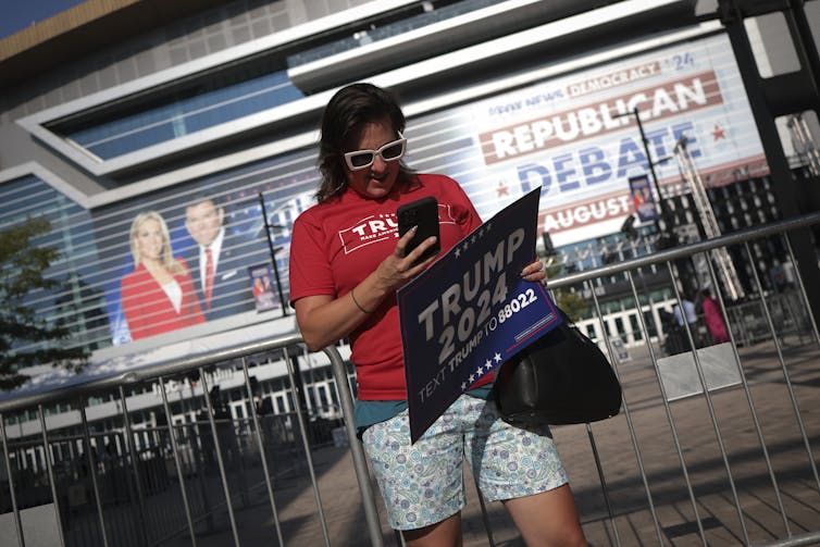 A dark-haired person in sunglasses and a red shirt holding a Trump sign in front of a large building.