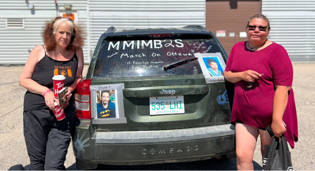 Two women stand next tot he back of a dark Jeep with a photo of a man and young boy taped it to it. MMIMB2S and March of Ottawa are also written on the car.