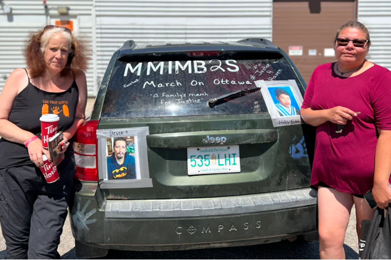 Two women stand next tot he back of a dark Jeep with a photo of a man and young boy taped it to it. MMIMB2S and March of Ottawa are also written on the car.