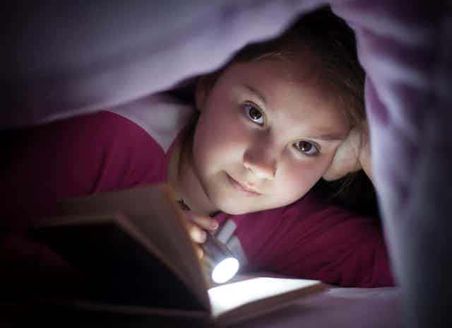 A young girl shines a flashlight on an open book while underneath her bed cover.