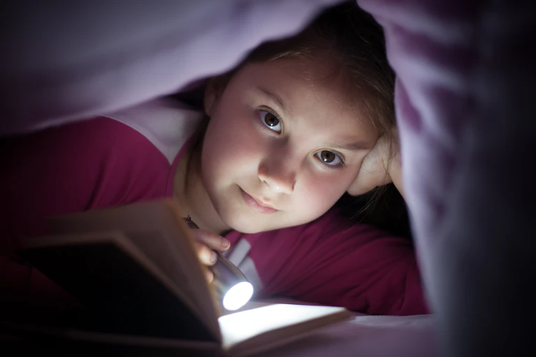 A young girl shines a flashlight on an open book while underneath her bed cover.