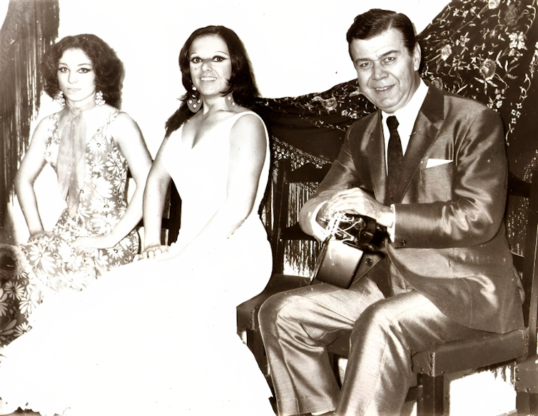Un hombre trajeado con una guitarra en la mano sonríe a cámara al lado de dos mujeres vestidas de flamencas que también sonríen.