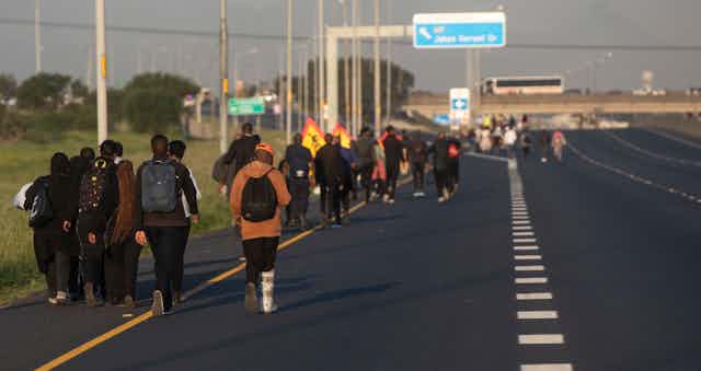 A highway with a long line of people of all ages walking next to the side of the road, some carrying bags.