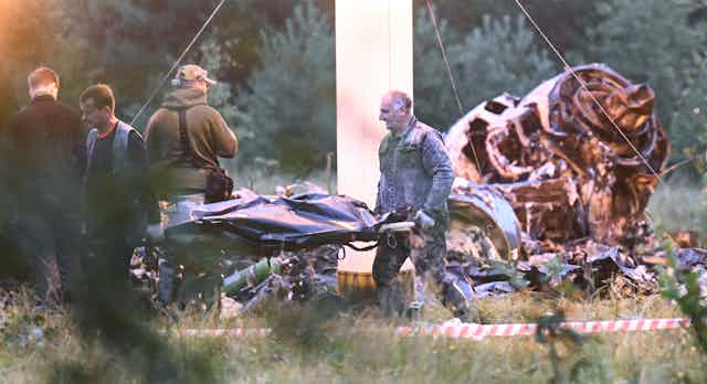 Workers carry a stretcher with a body bag. Scorched airplane wreckage is behind them.