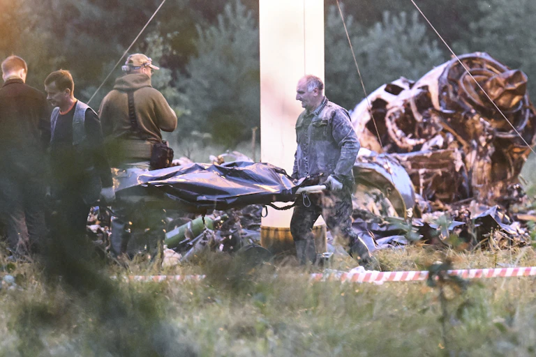 Workers carry a stretcher with a body bag. Scorched airplane wreckage is behind them.