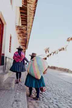 Dos mujeres caminan por una calle adoquinada vistiendo sombreros y ropas de colores.