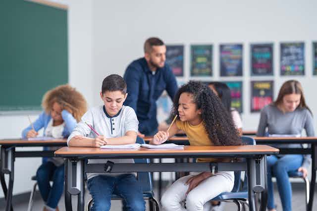 k 12 education research A male and female student work at a desk while a teacher helps other students in the classroom.