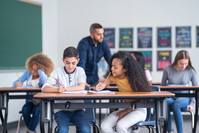A male and female student work at a desk while a teacher helps other students in the classroom.