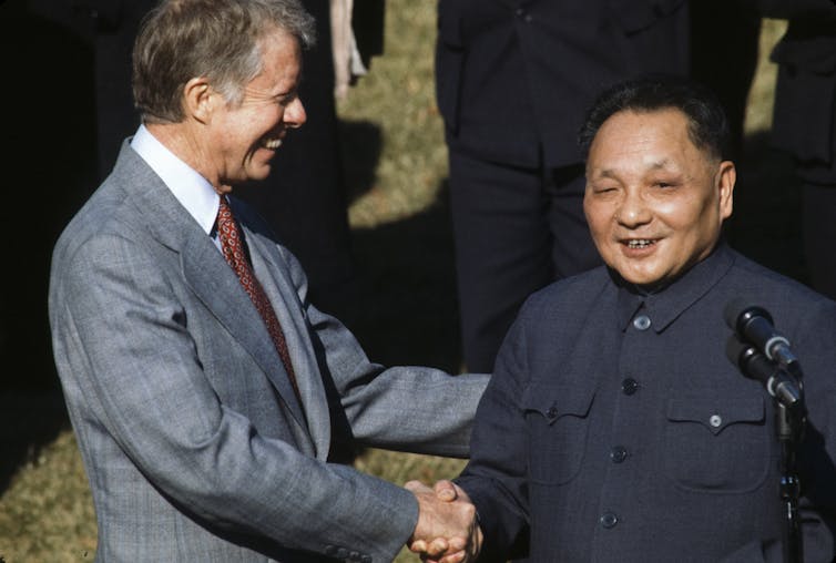 Two men, Jimmy Carter and Deng Xiao Ping, smile and shake hands while standing on a lawn.