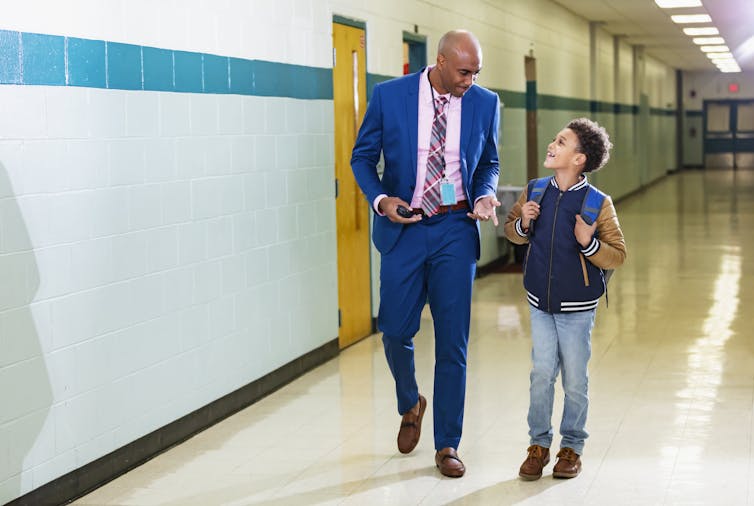 A man in a blue suit accompanies an elementary school-aged boy as they walk down a school hallway.