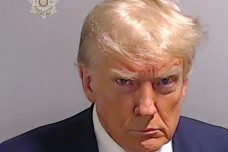 A man with blond hair, wearing a navy blue suit jacket with a burgundy tie and white shirt, stares at the camera with a grimace on his face