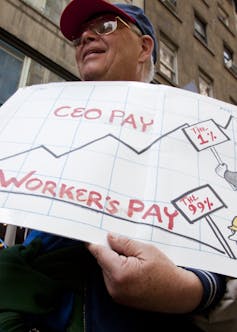 Man in cap with white sign printed with ceo pay: the 1% and worker's pay: the 99%.