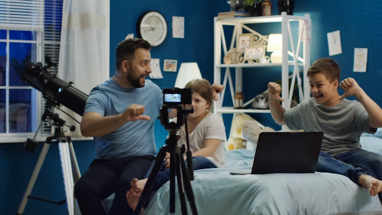 A man and two young children sit in front of cameras and a laptop.