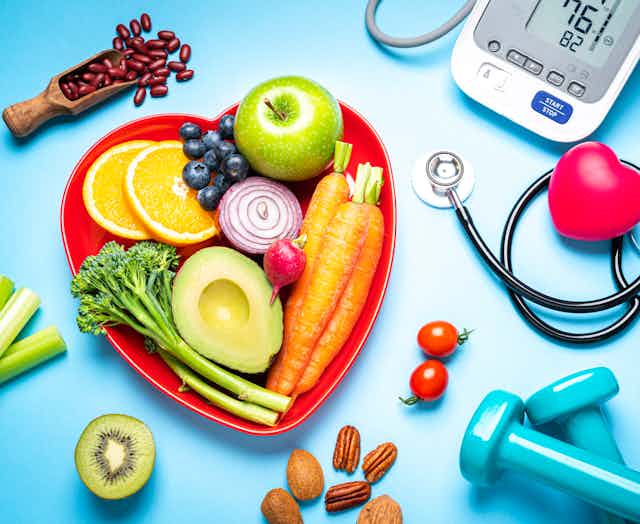 A red heart-shaped plate filled with fruits and vegetables sits next to dumbbells, a blood pressure reader and a stethoscope.