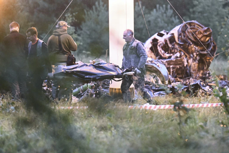 Des ouvriers transportent une civière avec un sac mortuaire. Derrière eux, des débris d'avion calcinés.