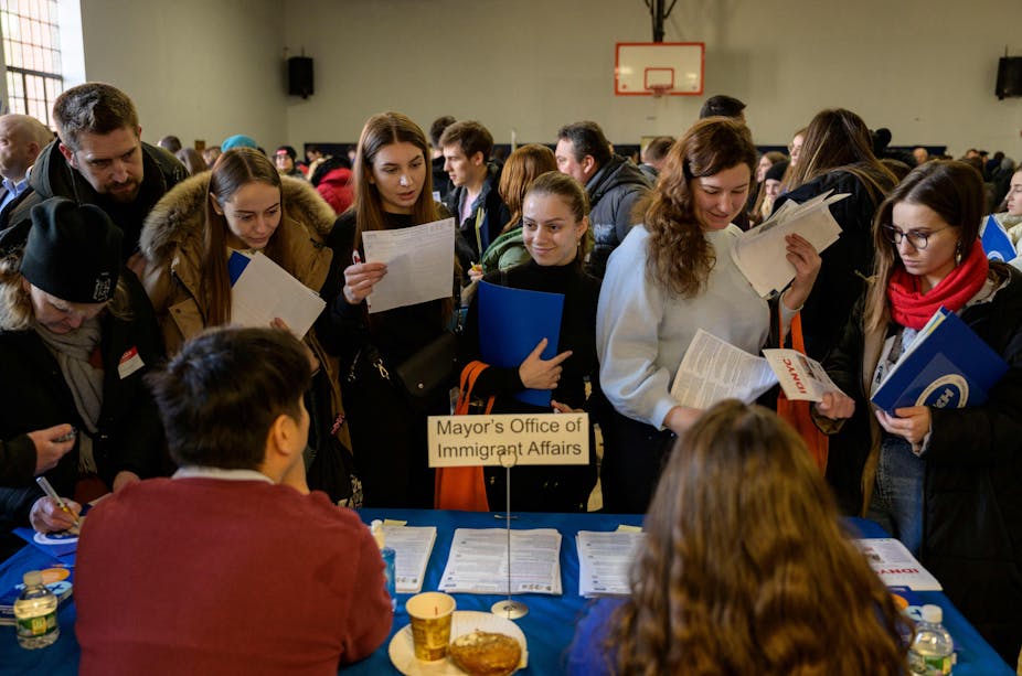 In a crowded room, people stand before an attended table with papers in their hands.