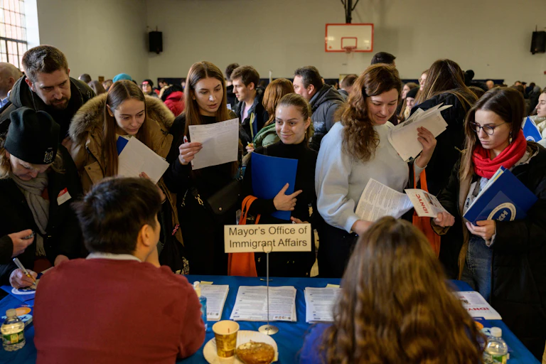 In a crowded room, people stand before an attended table with papers in their hands.