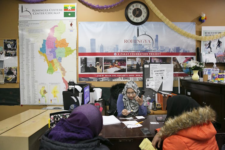 A woman wearing a hijab sits at a desk and looks down at a piece of paper in her hand. Two people wearing winter coats sit across from her with their backs to the camera.