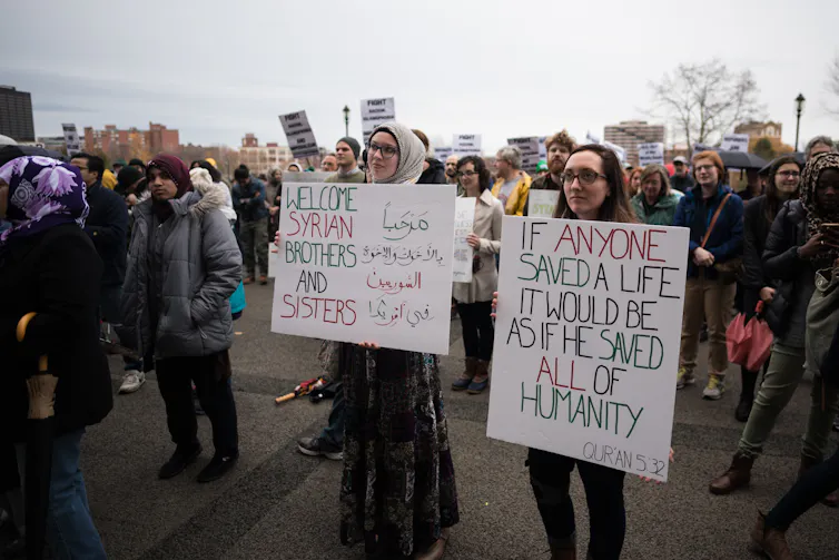 Two women stand on a street holding large signs while people stand behind them.