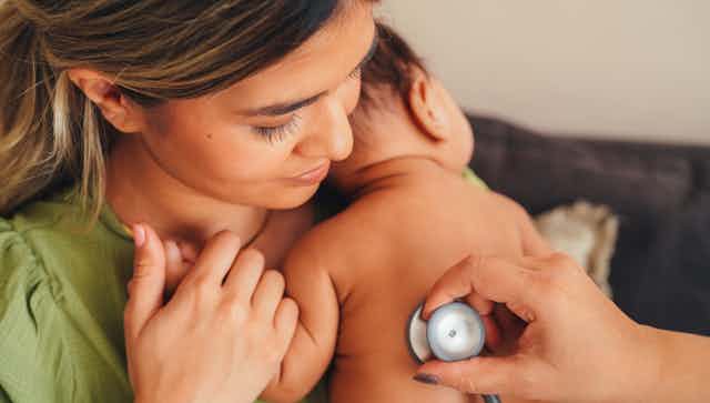 Mother holds a young baby to her chest as a doctor places a stethoscope on its back. 