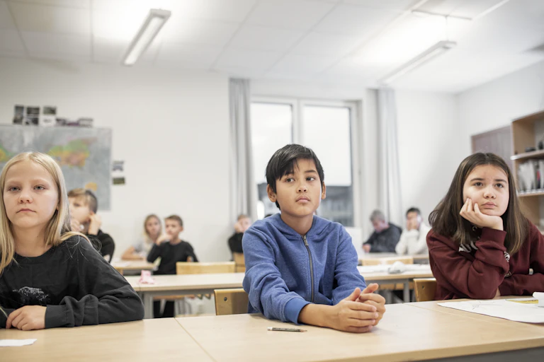 Three kids -- two girls and a boy -- sit at a desk looking pensive.