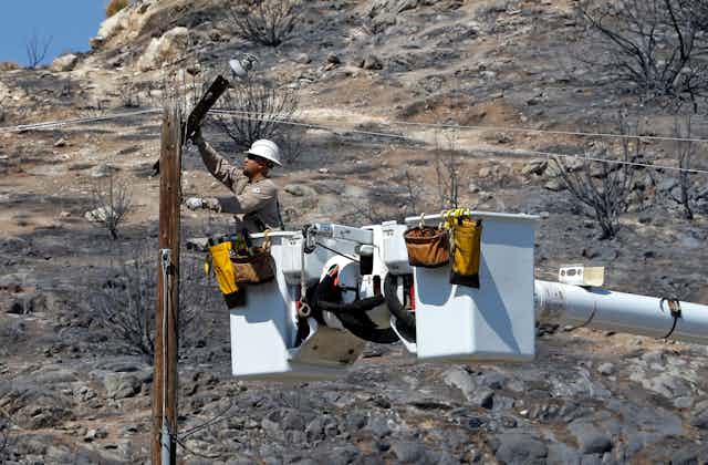 A lineman in a bucket truck bucket repairs a burned power line with a completely charred landscape behind him.