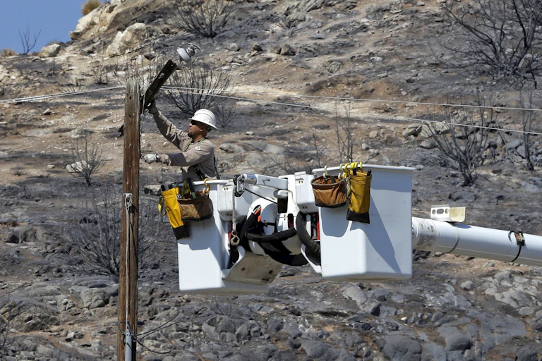 A lineman in a bucket truck bucket repairs a burned power line with a completely charred landscape behind him.