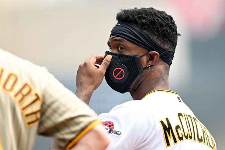 A baseball player standing beside the field adjusts a black mask over his face.